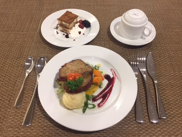 A plated meal on a woven placemat featuring a main dish with a slice of meat, mashed potatoes, vegetables, and a decorative sauce. Next to it is a dessert plate with a piece of layered cake topped with cocoa powder and garnished with whipped cream and berries. There is also a white cup and saucer set placed upside down. Silverware including two spoons on the left and a fork, knife, and spoon on the right are arranged around the plates.
