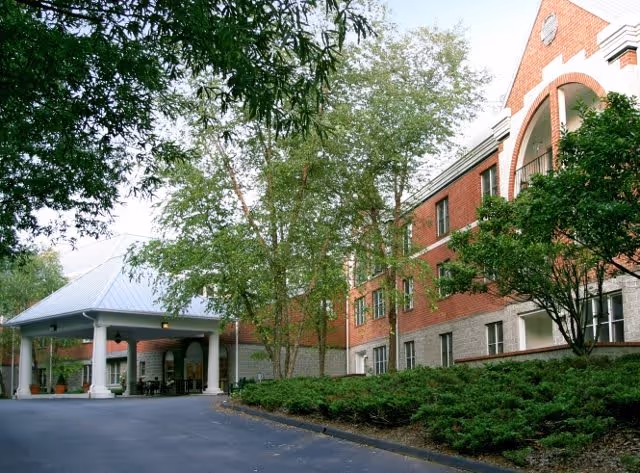 Exterior view of a senior living facility named Signal Mountain Senior Living, showing a brick building with white trim, a covered entrance supported by white columns, surrounded by trees and greenery.