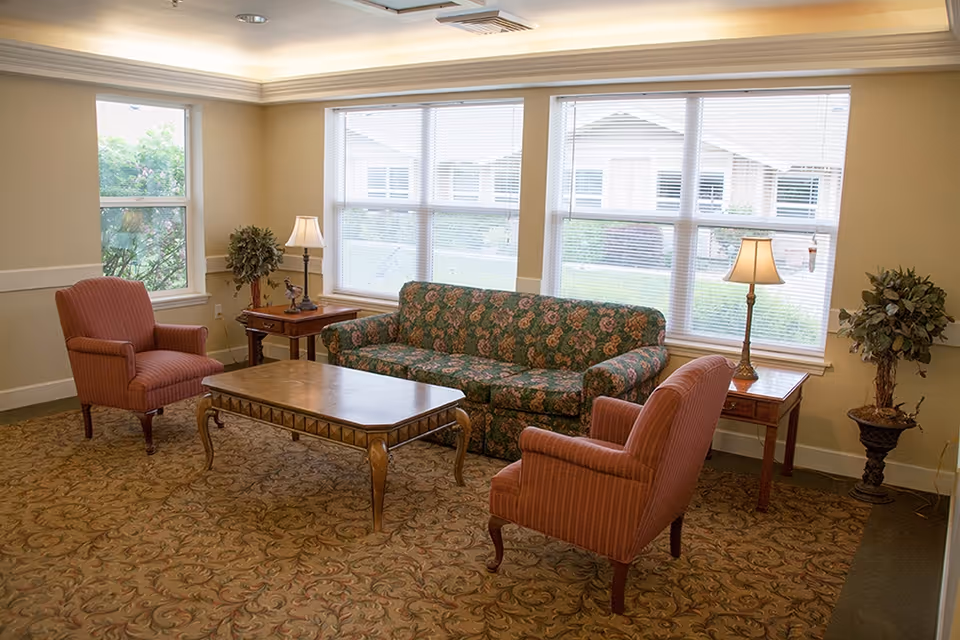 Sunlit common sitting room with a floral sofa, two upholstered armchairs, a wooden coffee table and side tables beneath large windows.