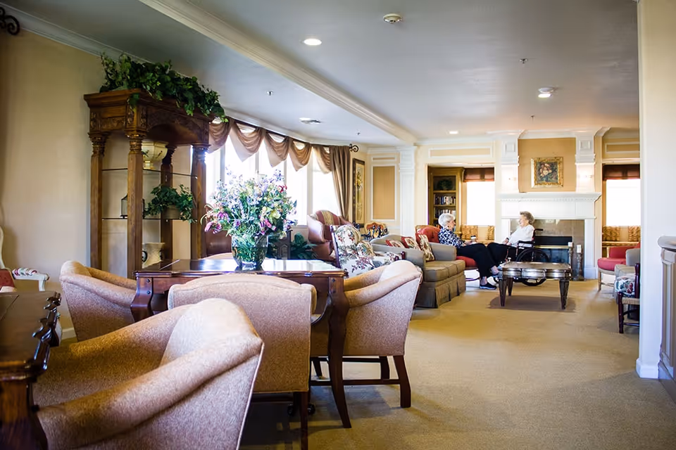 A spacious and well-lit living room area in a senior living facility with comfortable upholstered chairs and sofas arranged around a coffee table. Two elderly women are seated and engaged in conversation near a fireplace. The room features large windows with draped curtains, decorative plants, and framed artwork on the walls.