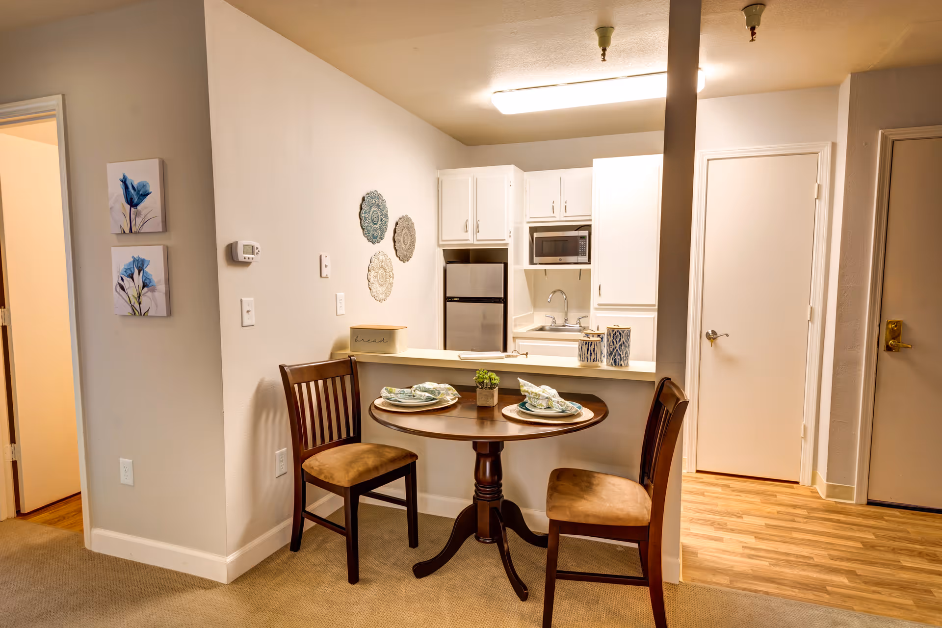 Small dining area with a round table set for two in front of a compact kitchenette inside an apartment.