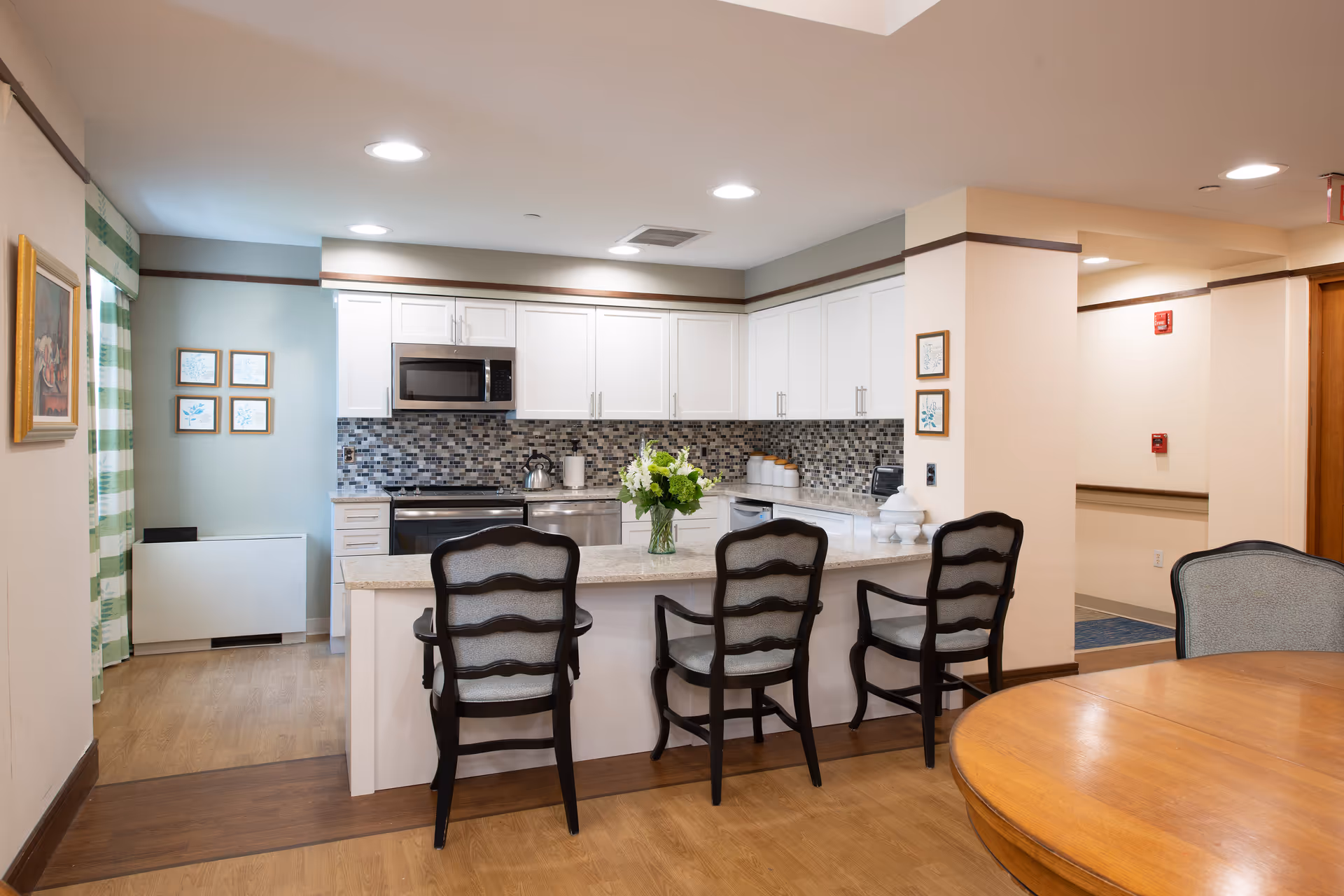 A modern kitchen area in a senior living facility with white cabinets, a mosaic tile backsplash, and stainless steel appliances including a microwave and stove. There is a countertop with three cushioned chairs and a vase of flowers on it. To the right, part of a wooden dining table with a chair is visible. The room has recessed lighting and light wood flooring.