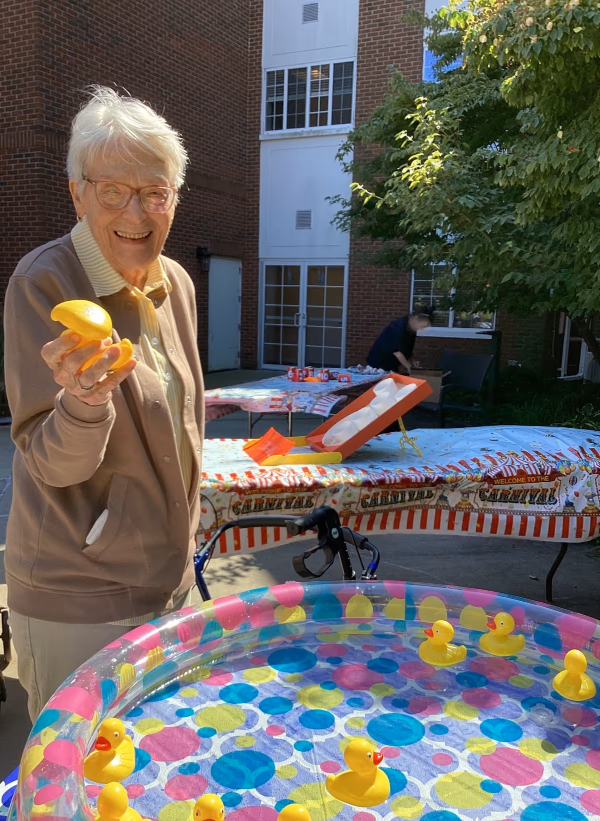 An elderly woman smiling and holding a yellow rubber duck near a colorful inflatable pool filled with water and several yellow rubber ducks. Behind her, there are tables covered with carnival-themed tablecloths and various items, set outdoors near a brick building with windows and a tree providing shade.