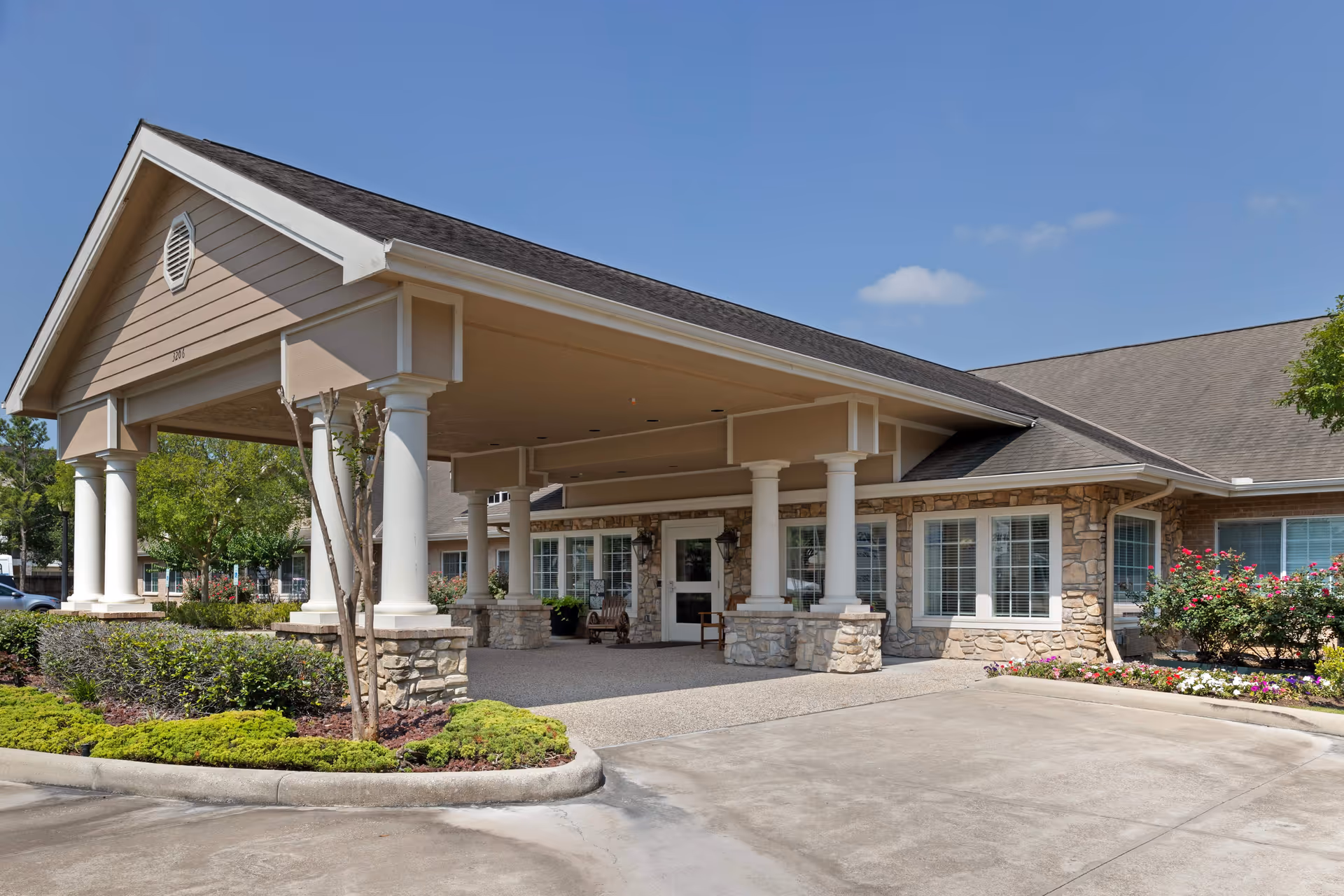 Exterior view of Cinco Ranch Alzheimer's Special Care Center showing the entrance with a covered driveway supported by white columns, stone facade, large windows, and landscaped greenery under a clear blue sky.