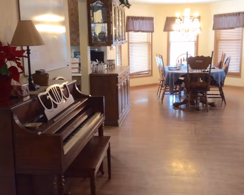 Interior room with an upright piano in the foreground and a dining table with chairs under a chandelier by windows.