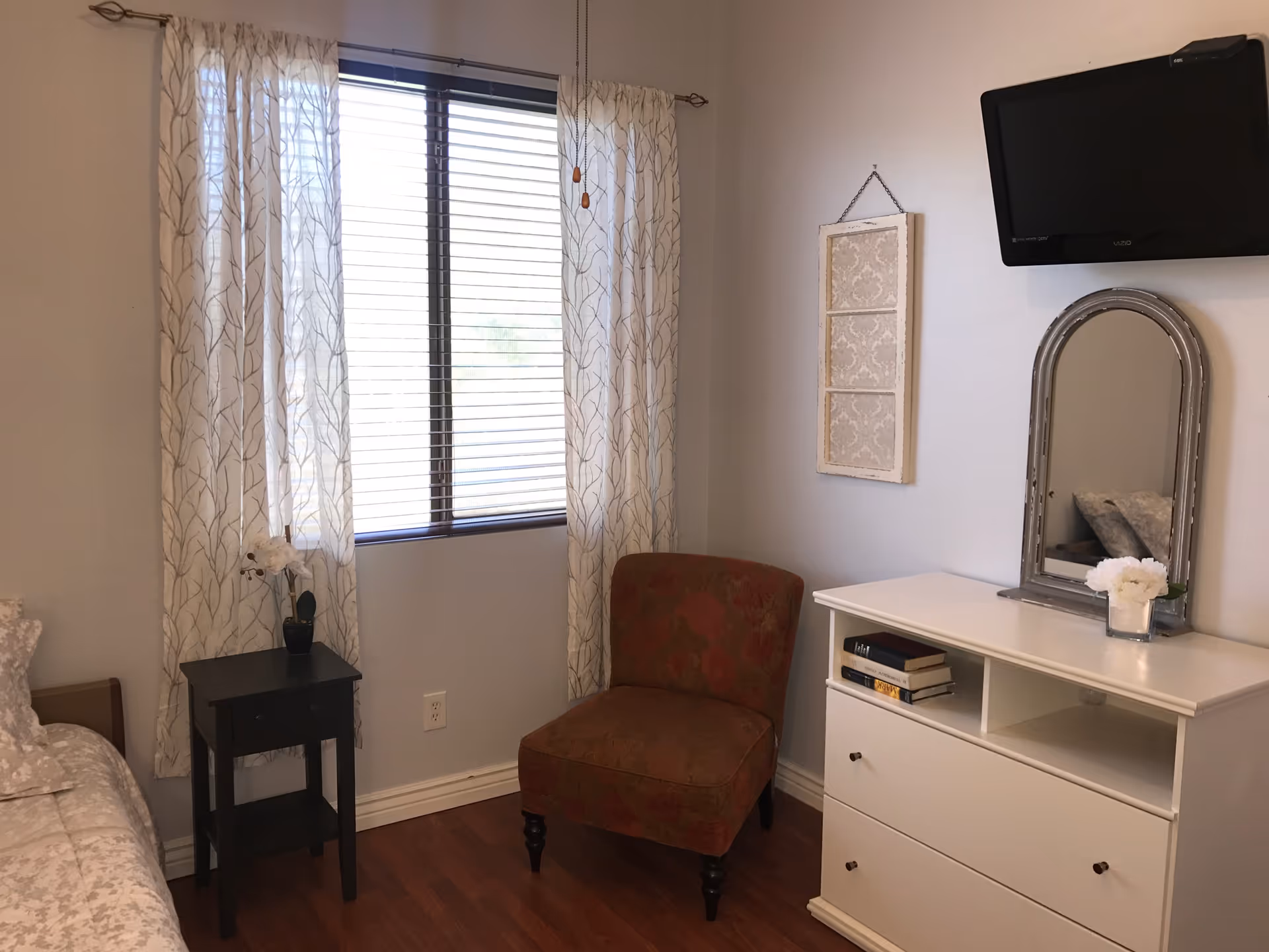 A bright bedroom with a window and patterned curtains, an upholstered chair, small nightstand, and a white dresser with mirror and wall-mounted TV.
