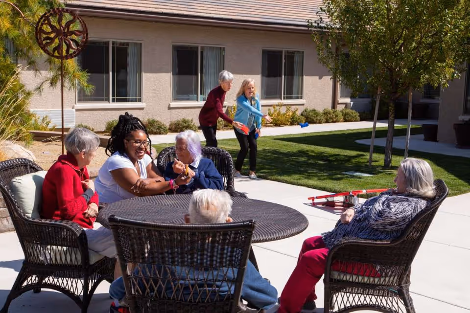 A group of elderly people and a caregiver sitting and chatting around a round outdoor table with wicker chairs in a sunny courtyard. In the background, two women are playing a bean bag toss game on the grass near a building with multiple windows.