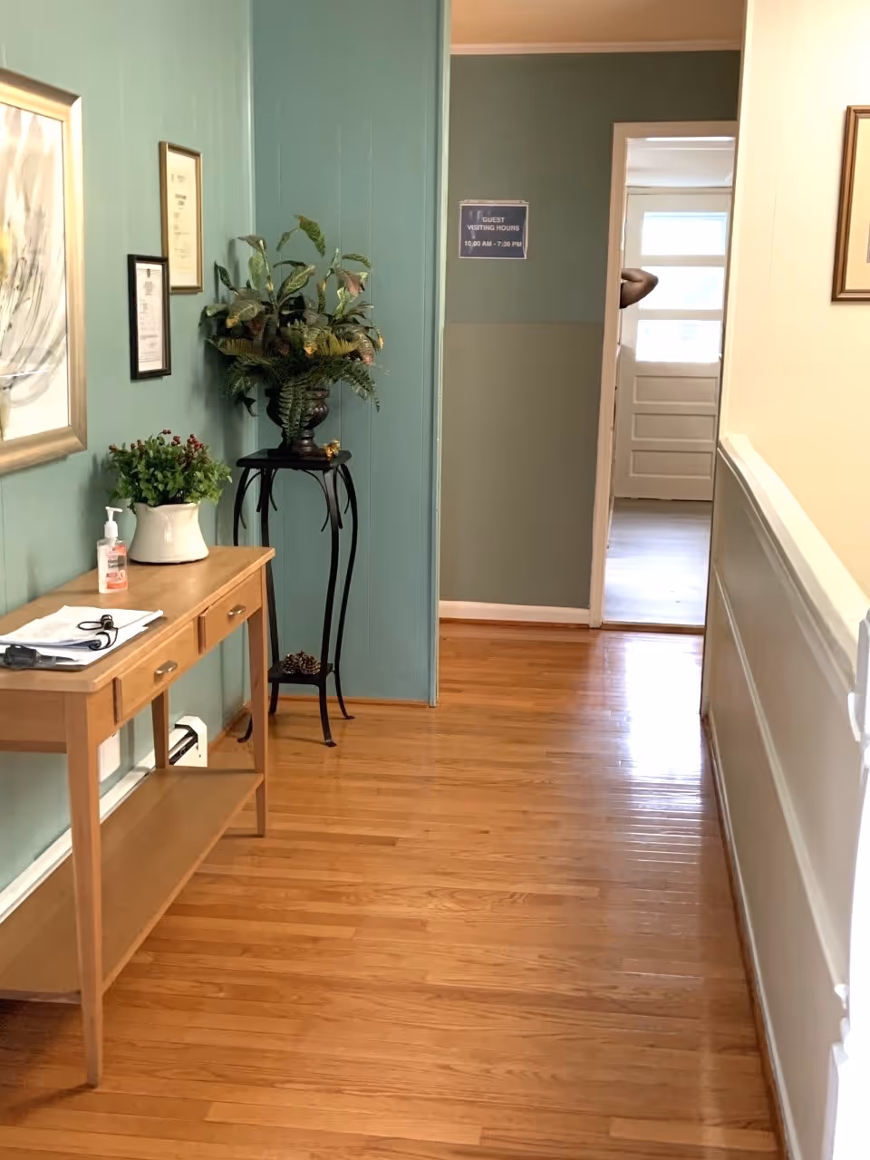 A hallway with wooden flooring and light green walls. On the left side, there is a wooden console table with a white vase containing green plants, a bottle of hand sanitizer, and some papers. Above the table, there are framed pictures on the wall. Next to the table, there is a tall black plant stand with a large green leafy plant. At the end of the hallway, there is an open doorway leading to a room with a white door and windows. A sign on the wall near the doorway reads 'Guest Visiting Hours 10:00 AM - 7:30 PM'.