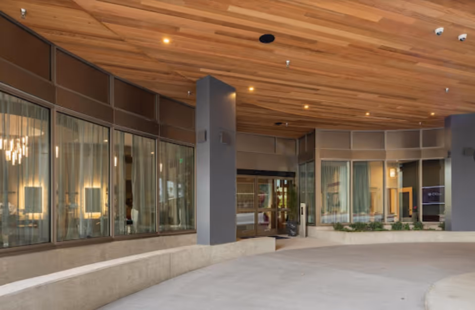 Covered entrance area of a building with large glass windows and doors, a wooden ceiling with recessed lighting, and concrete seating along the perimeter.
