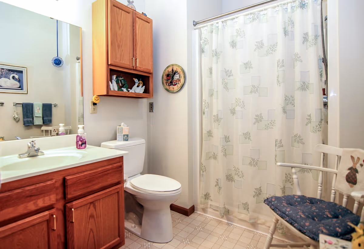 Bright bathroom featuring a wooden vanity and sink, toilet, shower with patterned curtain, wall cabinet and a cushioned chair.