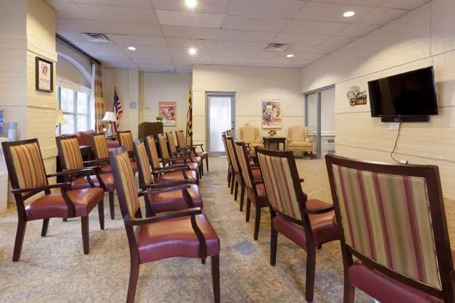 A meeting or activity room with rows of wooden chairs featuring striped upholstery and red seats, arranged facing a wall-mounted flat screen TV. The room has beige walls, carpeted floor, and ceiling lights. There are two armchairs and a small table in the back corner, along with framed posters and an American flag near a window with curtains.
