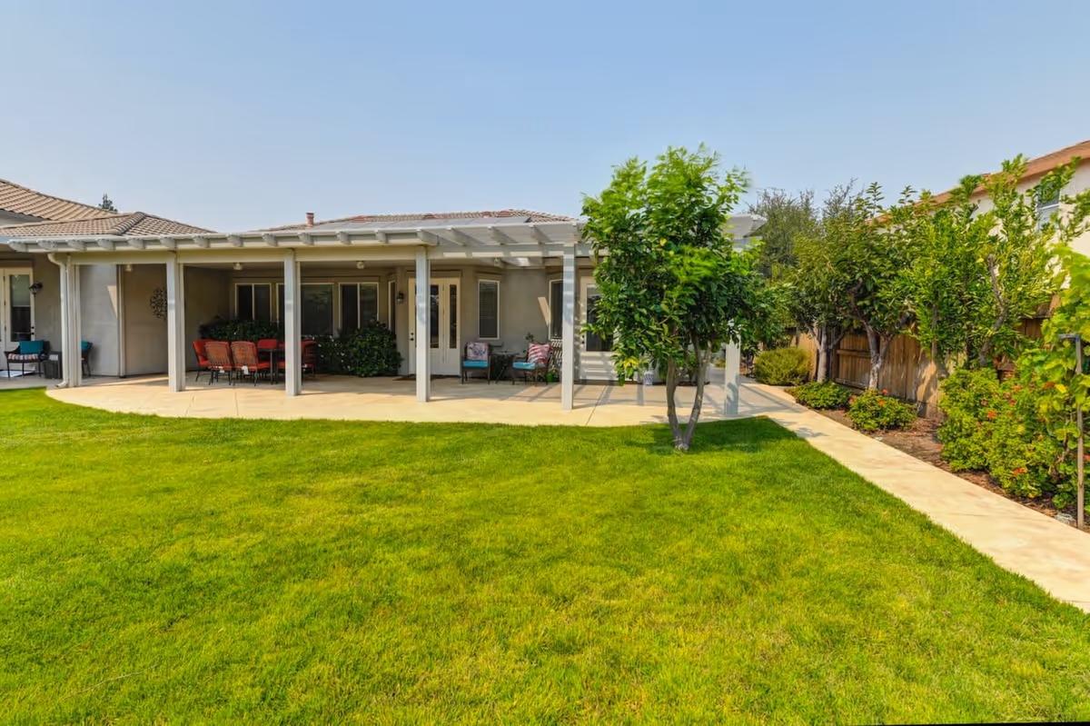 A backyard view of a single-story building with a covered patio area featuring outdoor seating and a table. The patio is supported by white columns and overlooks a well-maintained green lawn with a concrete pathway and several trees and shrubs along the fence line.
