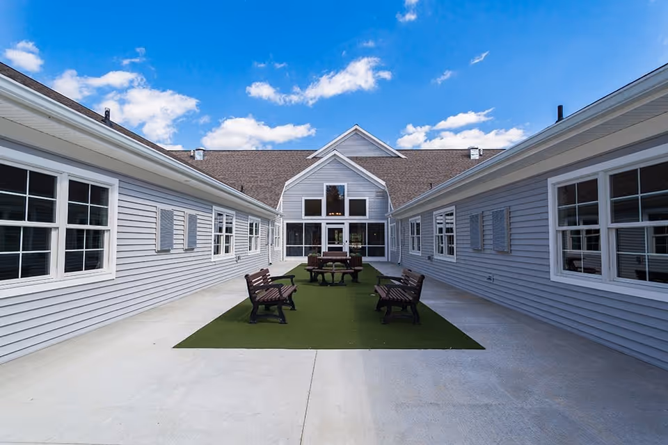 Outdoor courtyard area of a senior living facility with benches and a picnic table on a green artificial turf, surrounded by light gray buildings with white-trimmed windows under a blue sky with scattered clouds.
