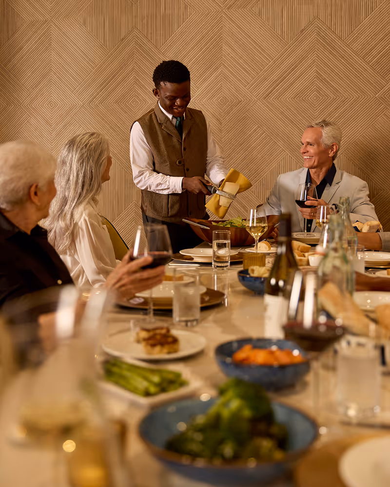 A young waiter serving salad to a group of elderly people seated around a dining table set with plates, glasses of wine, and various dishes in a warmly lit room with patterned wallpaper.