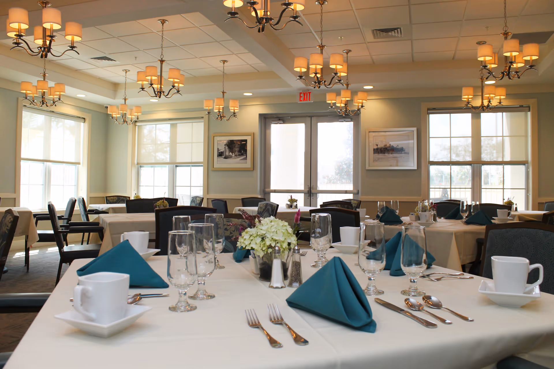 A bright and elegant dining room with tables set for a meal, featuring white tablecloths, teal folded napkins, glassware, white cups and saucers, silverware, and small floral centerpieces. The room has large windows letting in natural light, framed pictures on the walls, and multiple chandeliers hanging from the ceiling.