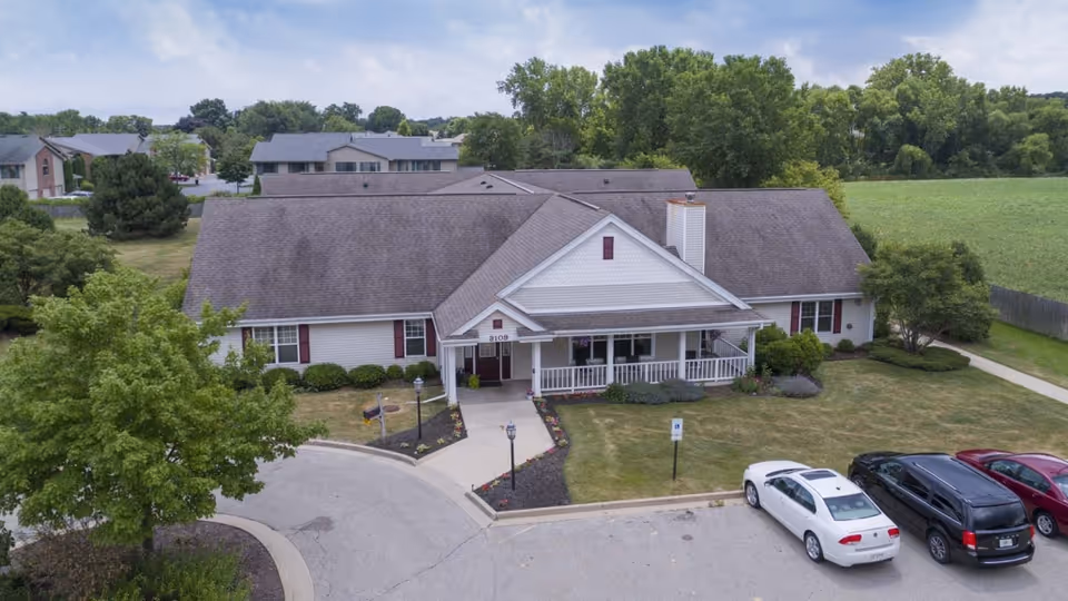 Aerial view of a single-story senior living facility building with a gray roof and white exterior walls. The building has a covered entrance with a small porch and is surrounded by a well-maintained lawn, shrubs, and trees. There is a parking lot in front with several parked cars, and neighboring buildings and green fields are visible in the background.