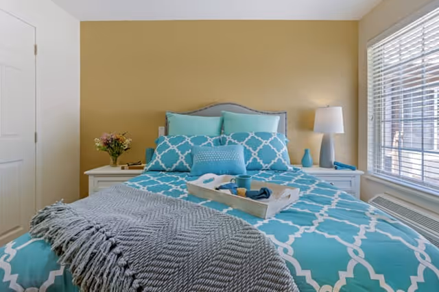 A neatly made bed with a turquoise and white patterned comforter and multiple pillows in shades of turquoise and light blue. A gray knitted throw blanket is draped over the foot of the bed. On the bed is a wooden tray holding a cup, a small bowl, and a rolled-up napkin. On either side of the bed are white nightstands, one with a vase of flowers and the other with a lamp and a small turquoise vase. A window with white blinds is on the right side of the room, and the walls are painted a warm beige color.