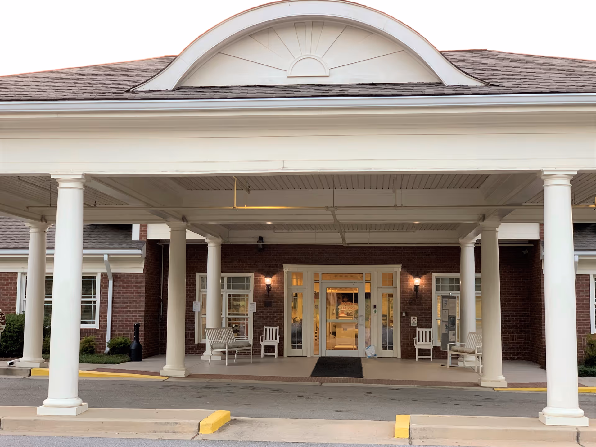 Covered porte-cochere with white columns and benches in front of a brick building's glass entry doors.