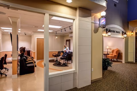 Interior corridor of a senior living facility showing a hair salon with styling chairs and a person attending a client and a nearby doorway labeled 'Music Hall'.