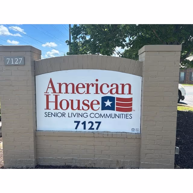 A brick signpost with a white sign displaying the text 'American House Senior Living Communities' along with a blue star and red stripes logo, and the number 7127. The sign is outdoors with trees and a parked car visible in the background.