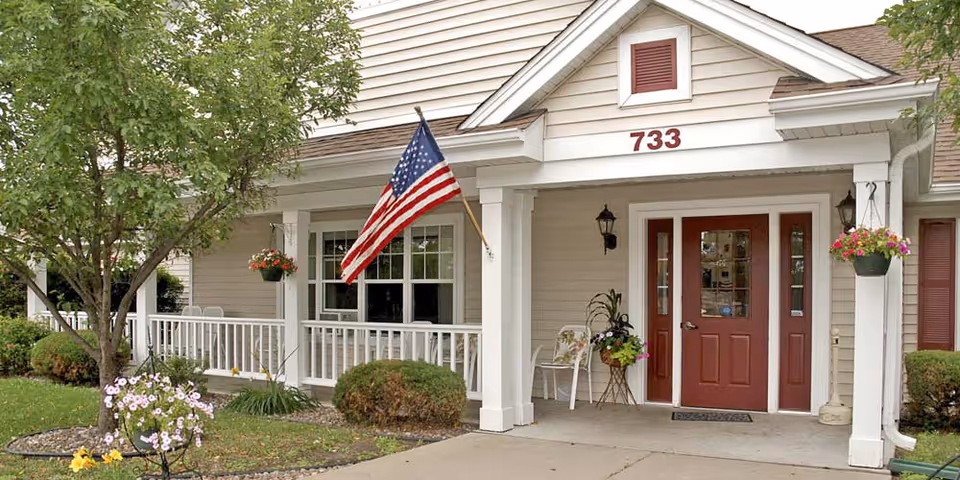 Front entrance of a senior living facility with beige siding, a red door, white columns, and an American flag mounted near the window. There are hanging flower pots and a small tree with flowers in the landscaped area near the sidewalk.