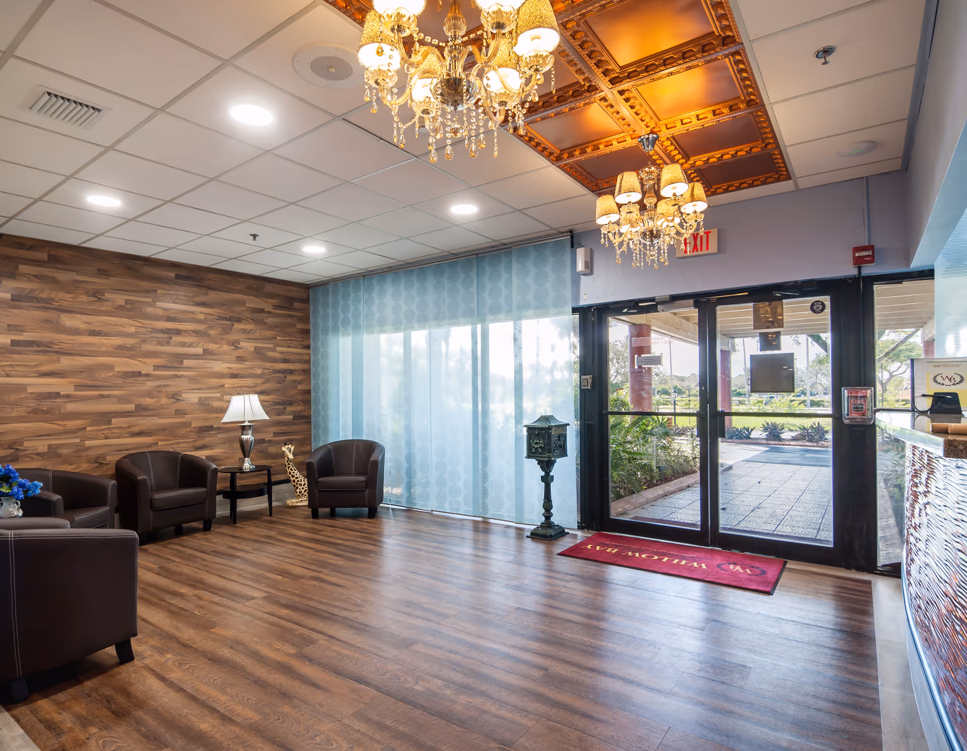 A bright and welcoming senior living facility lobby with wooden flooring and a wood-paneled accent wall. The space features several brown leather armchairs arranged around a small table with a lamp, a decorative giraffe statue, and two ornate chandeliers hanging from a ceiling with decorative panels. Large glass doors lead outside, with a red welcome mat that reads 'Willow Bay'.