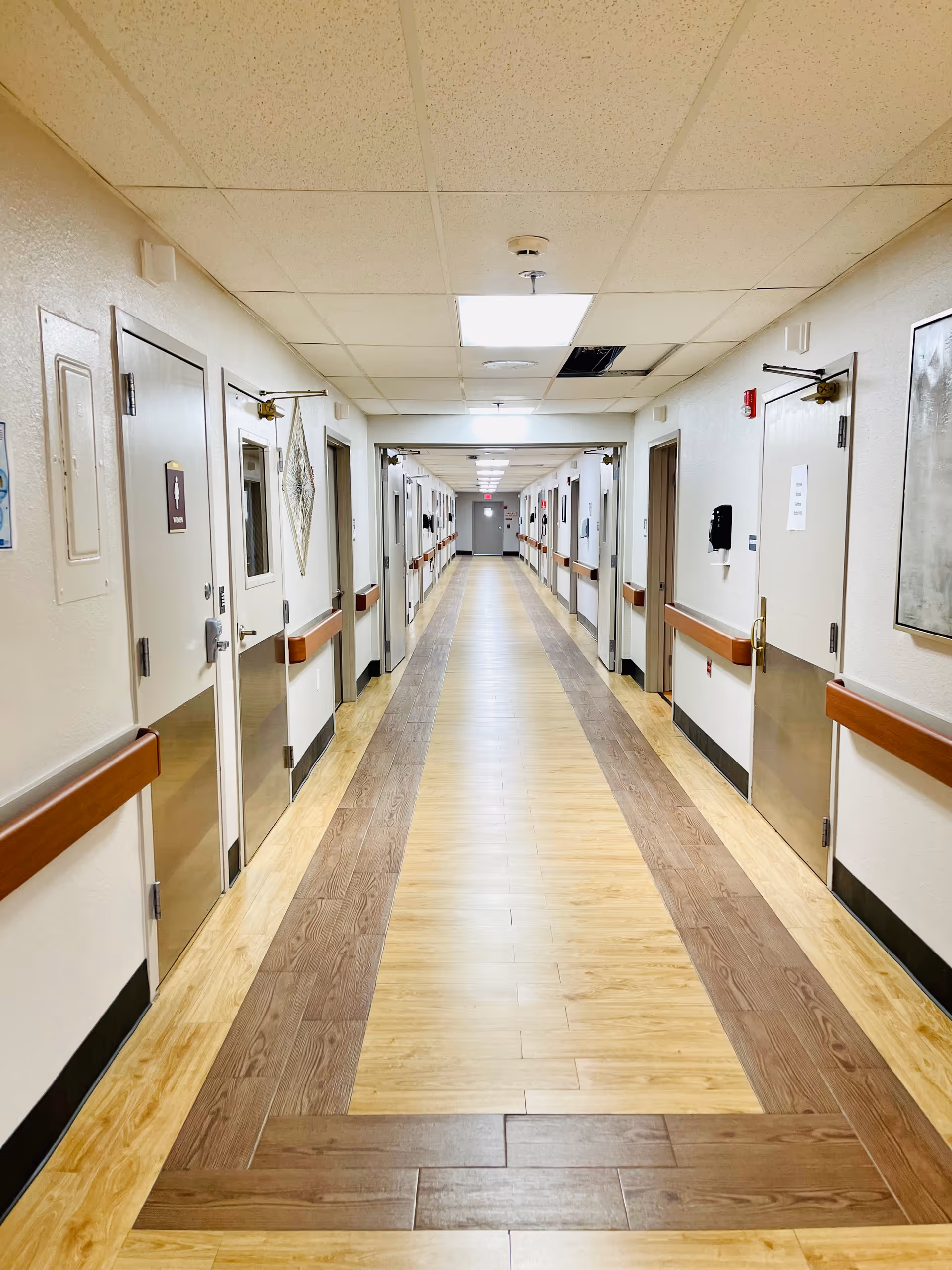 A long, well-lit hallway in a healthcare facility with light-colored walls and a patterned wooden floor. Doors line both sides of the corridor, some with signs and handrails along the walls. The ceiling has recessed lighting and a few ceiling tiles are missing or displaced.