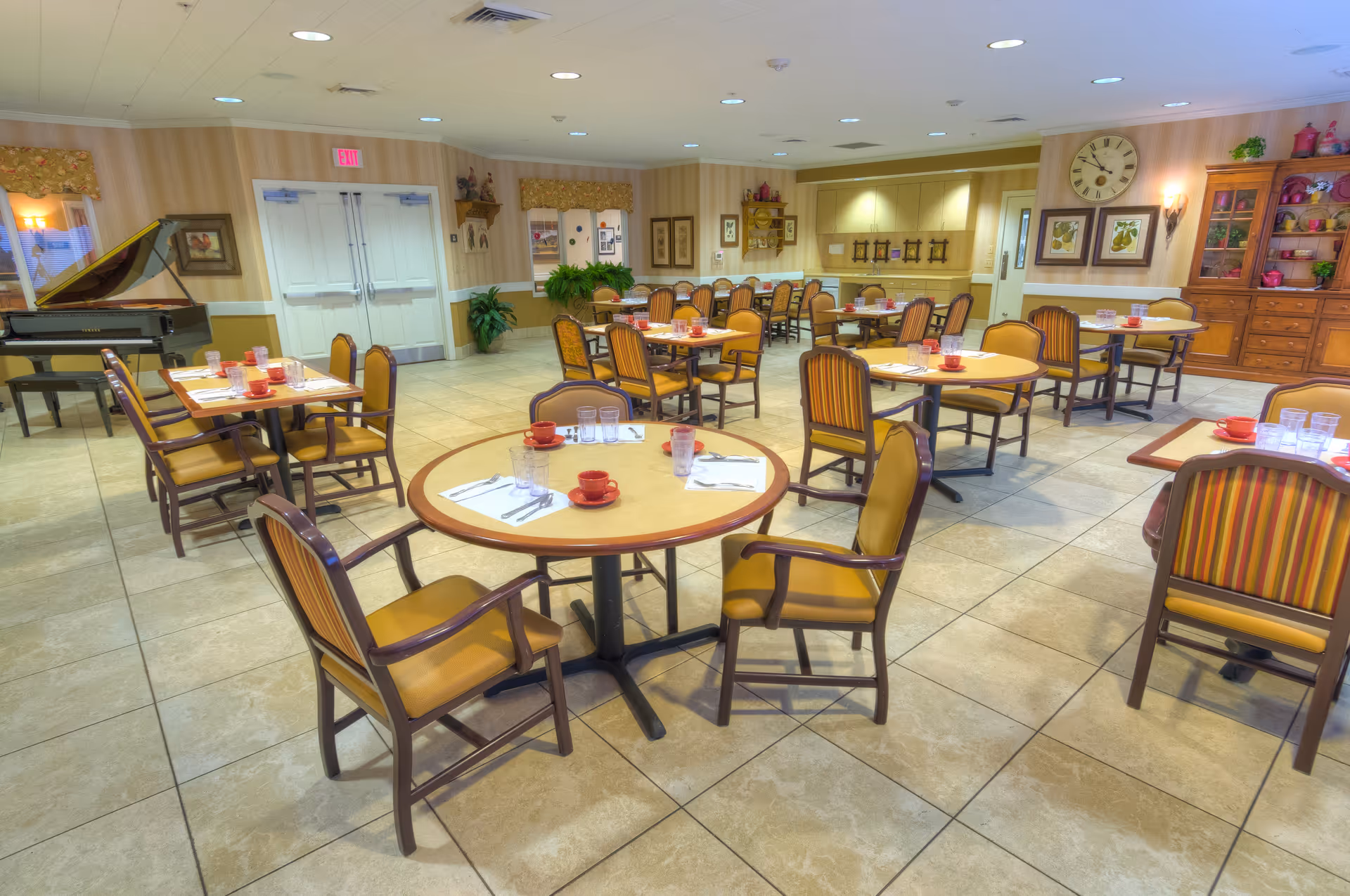 A spacious dining room with multiple round and rectangular tables set with red cups, glasses, and napkins. The room features beige tiled floors, yellow cushioned chairs with wooden frames, a large clock on the wall, framed artwork, a wooden cabinet with decorative items, and a grand piano in the corner. The walls are light-colored with a chair rail and some plants are placed around the room.