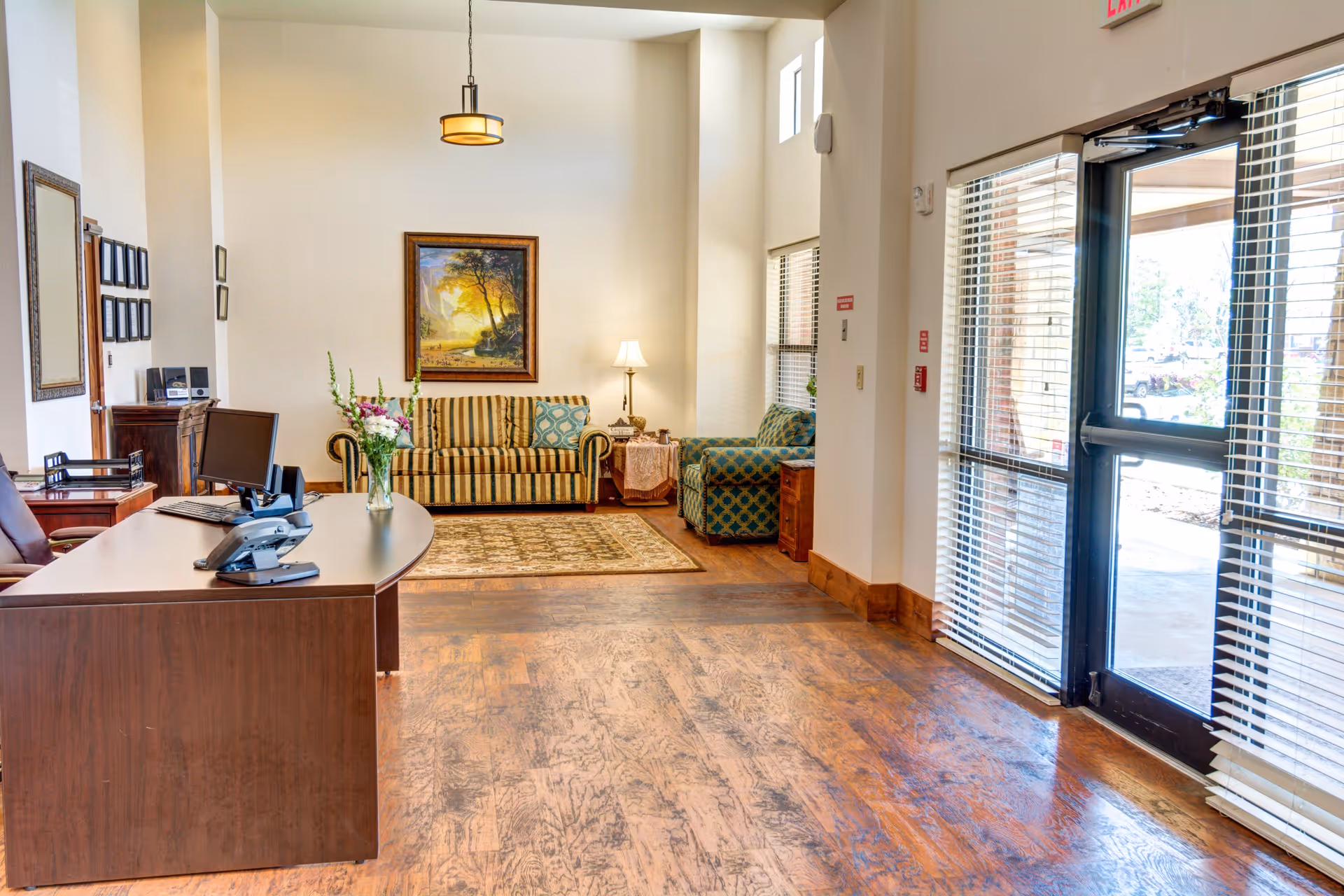 Reception/lobby area with a desk in the foreground, a striped sofa and armchair, framed art, and glass entry doors to the right.