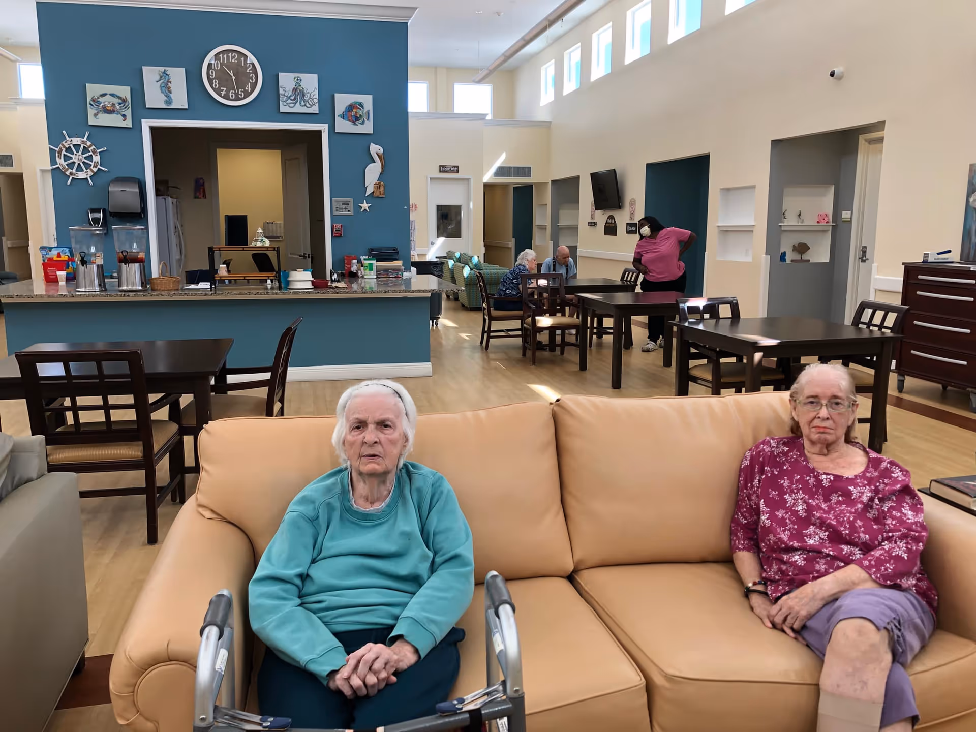 Two elderly women sitting on a beige couch in a senior living facility common area. Behind them, there are tables and chairs, a counter with coffee dispensers, and a blue accent wall decorated with nautical-themed artwork and a clock. Other residents and a staff member wearing a mask are visible in the background.