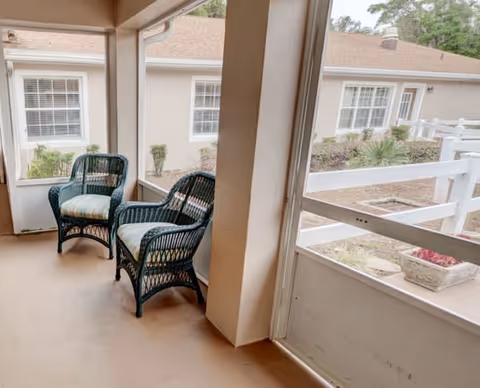 A screened-in porch area with two wicker chairs that have striped cushions. The porch overlooks a courtyard with plants and a white fence, and beige buildings with white-framed windows are visible in the background.