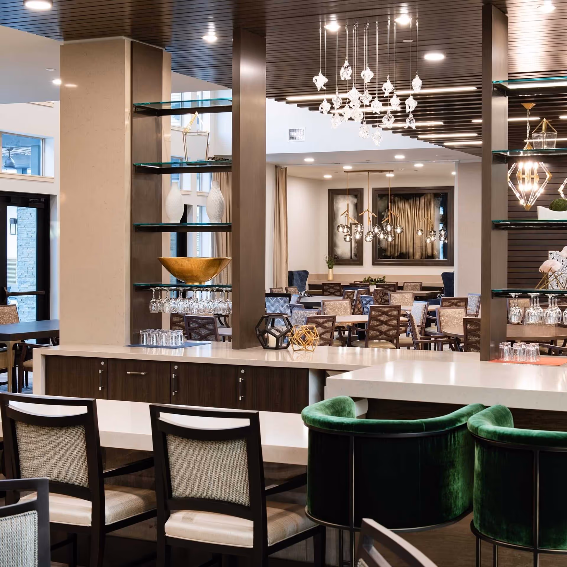 A modern dining area in a senior living facility featuring multiple tables and chairs arranged neatly. The space includes a bar counter with green velvet bar stools, glass shelves with decorative items, and contemporary hanging light fixtures. Large windows and a high ceiling add to the open and airy atmosphere.