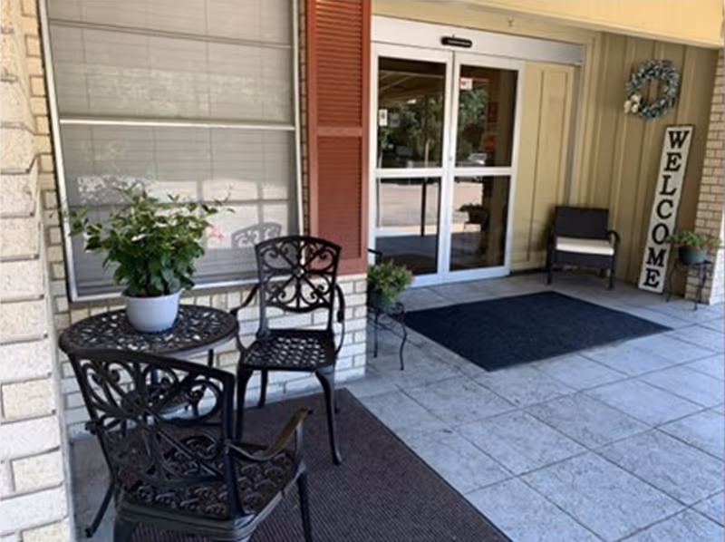 Covered entrance with glass double doors, a "WELCOME" sign, potted plants and outdoor seating.