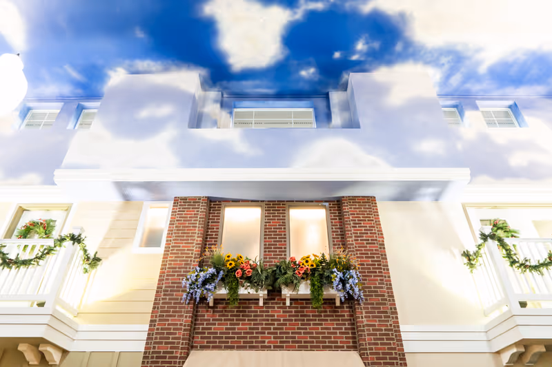 Exterior view of a building facade with a brick section featuring three windows adorned with flower boxes containing colorful flowers. The building has white balconies decorated with green garlands and a bright blue sky with clouds above.