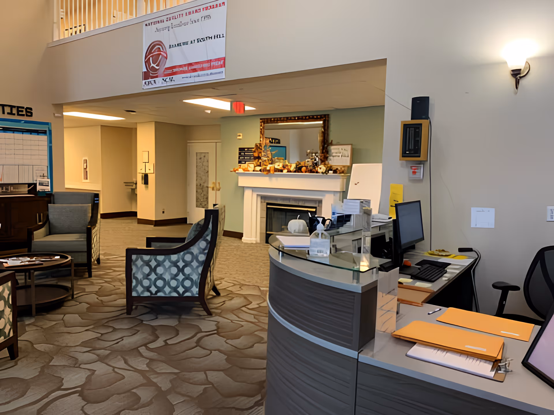 Reception area of Avamere at South Hill facility featuring a curved front desk with office supplies and computer, seating area with patterned armchairs and a round table, a fireplace with decorative items and a large mirror above it, and a hallway leading to other rooms.