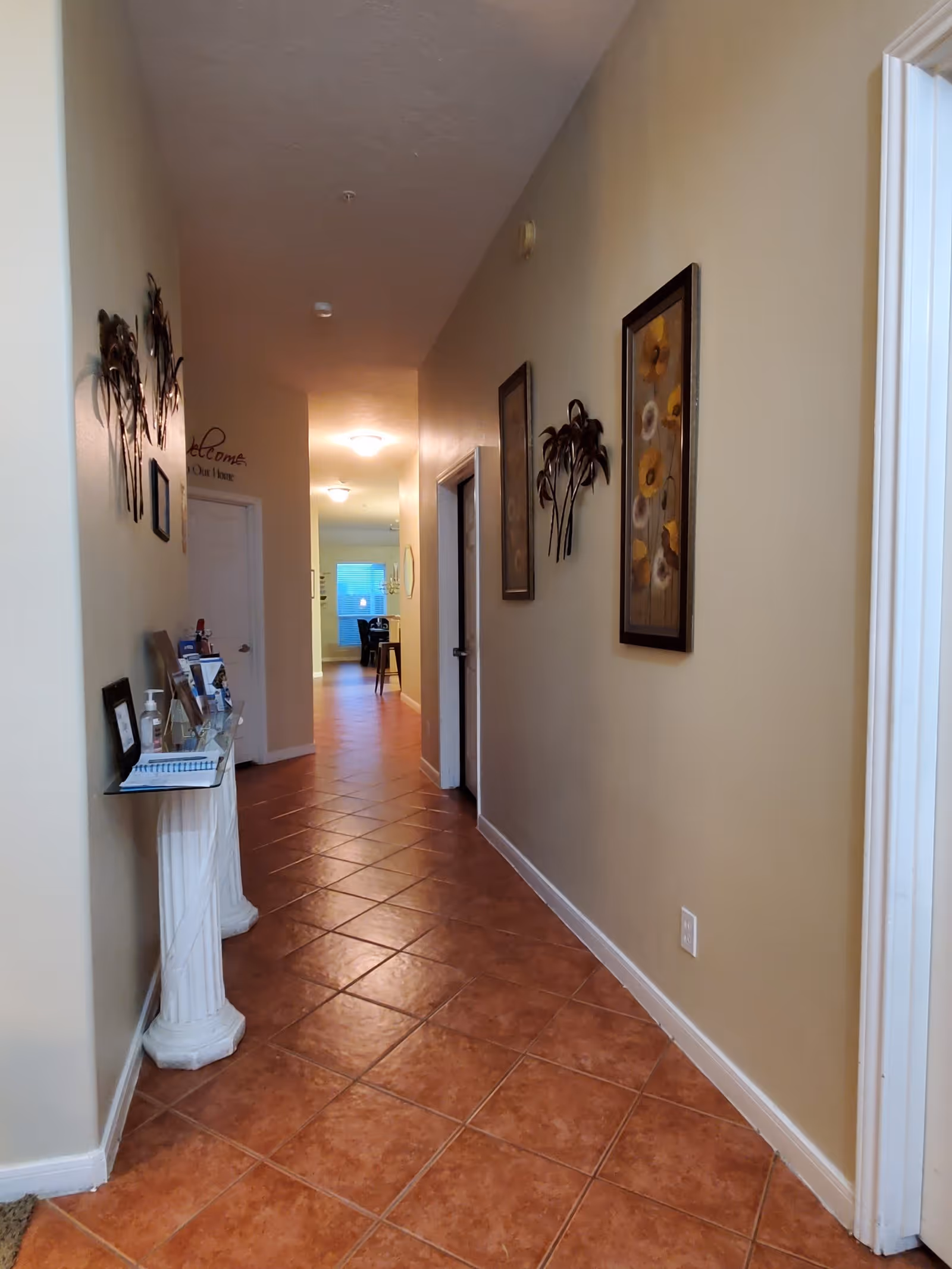 A long indoor hallway with terracotta tiled floor and beige walls. The hallway is decorated with framed floral artwork and metal wall art. There is a small glass table on a white pedestal holding various items on the left side. The hallway leads to a room with a window and dining table visible in the distance.