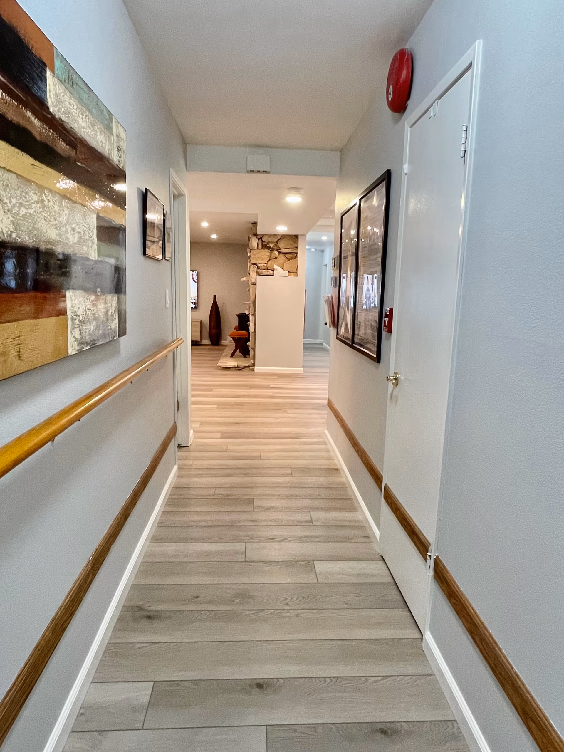 A well-lit hallway with light gray walls and wood flooring. The hallway has wooden handrails on both sides and framed artwork hanging on the walls. At the end of the hallway, there is a partial view of a room with a stone fireplace and decorative vases.