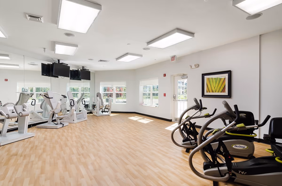 A bright exercise room with light wood flooring and white walls. The room contains several pieces of fitness equipment including NuStep machines and other exercise machines. Large mirrors cover one wall, and there are windows letting in natural light. A framed picture hangs on one wall near an exit door.