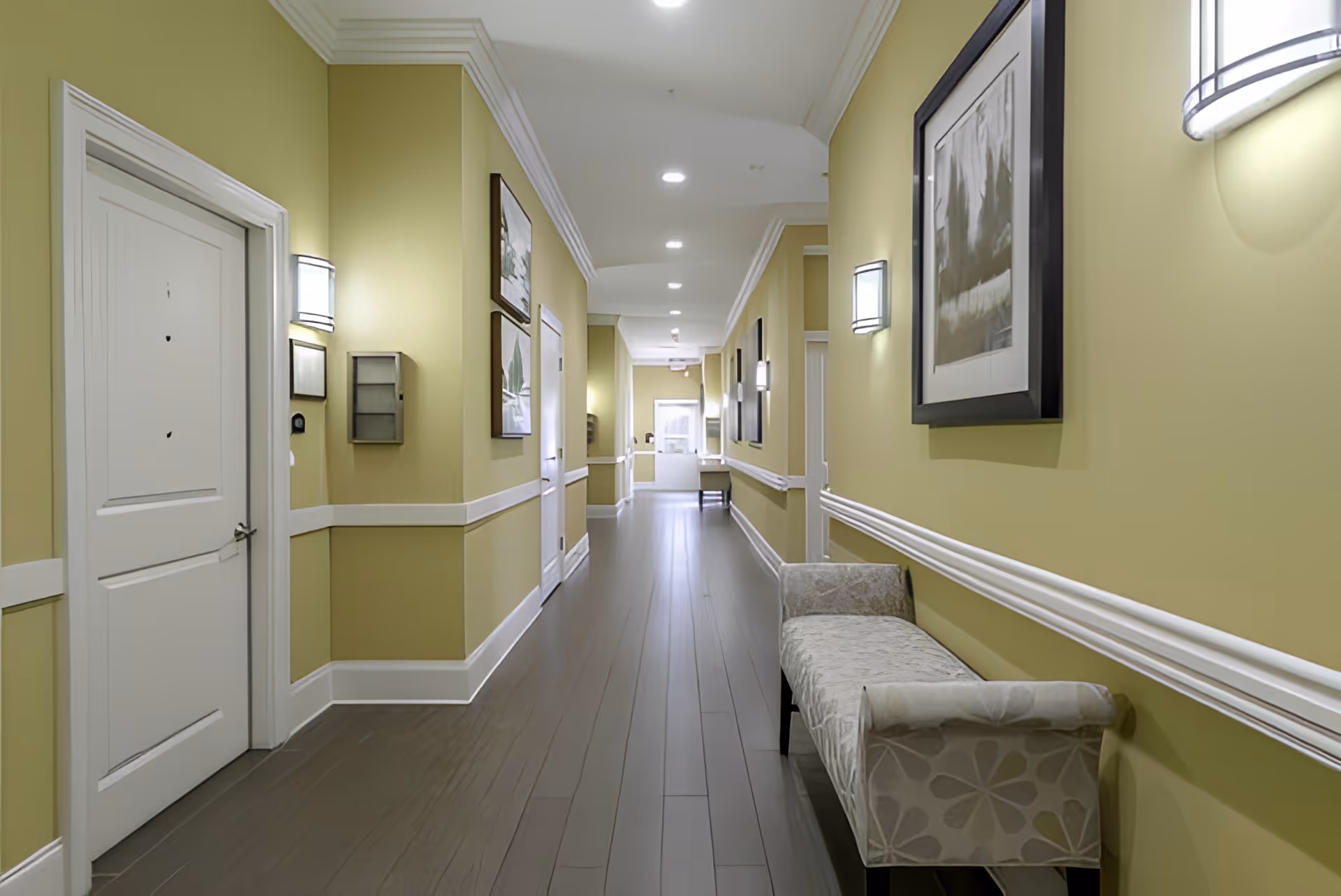 A well-lit hallway with yellow walls, featuring framed artwork, a bench, and doors leading to rooms.