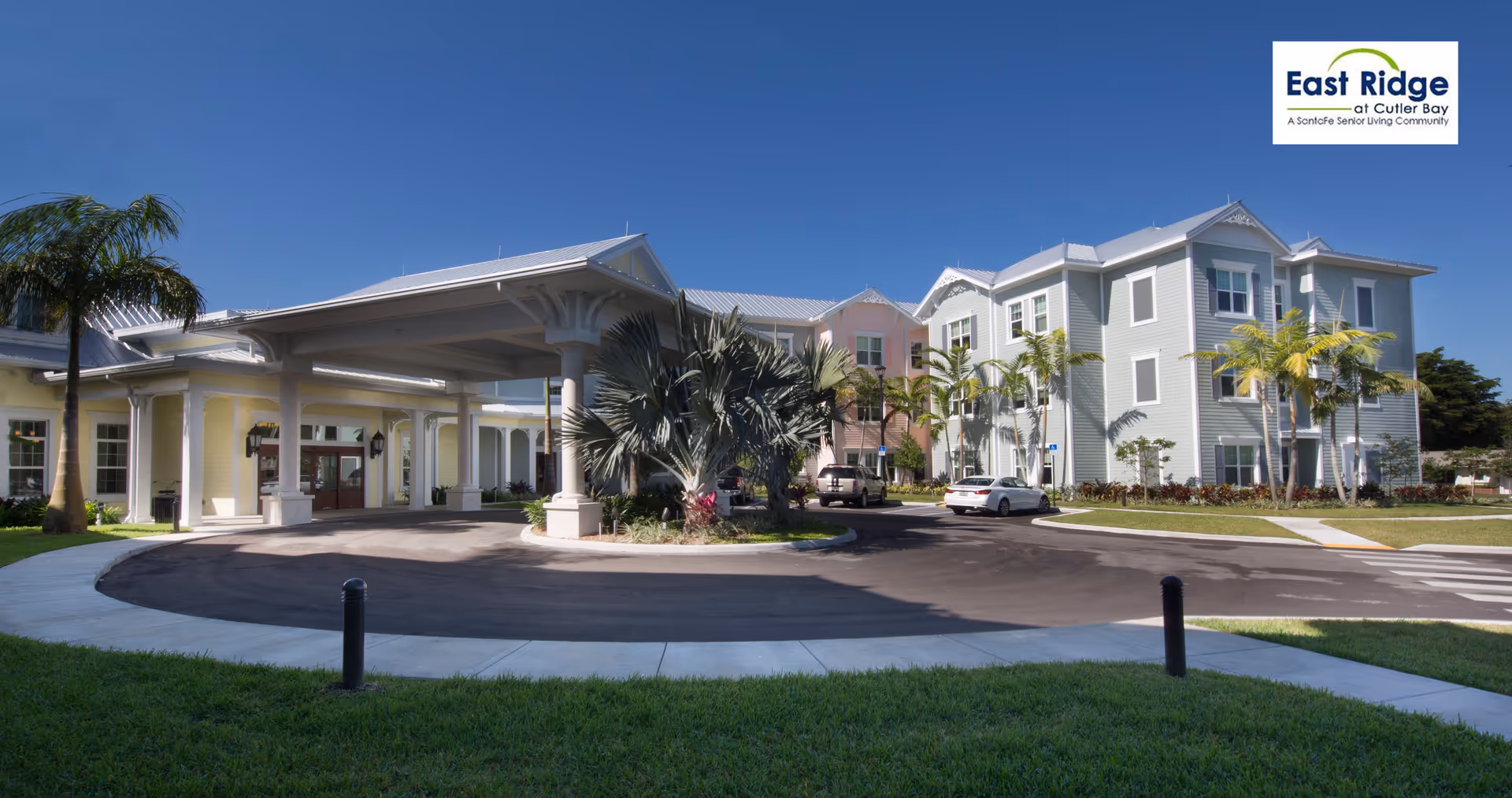 Exterior view of East Ridge Retirement Village, a senior living community, showing a large building with pastel-colored walls and a covered entrance driveway surrounded by palm trees and landscaping under a clear blue sky.