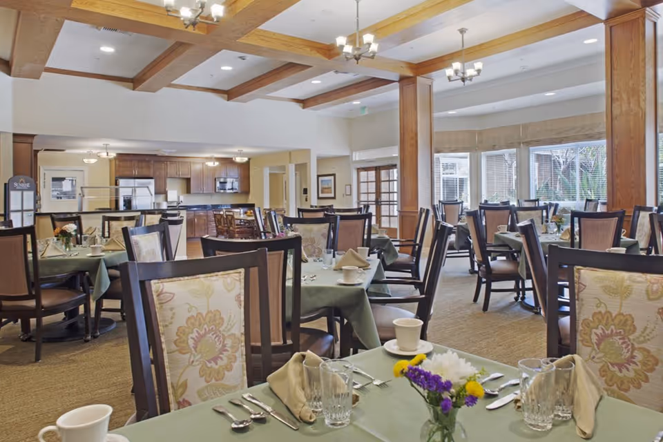 A dining room with multiple tables covered in green tablecloths, each set with cups, glasses, silverware, and napkins. The chairs have floral-patterned cushions. The room features wooden beams on the ceiling, large windows with blinds, and a kitchen area in the background with wooden cabinets and appliances.