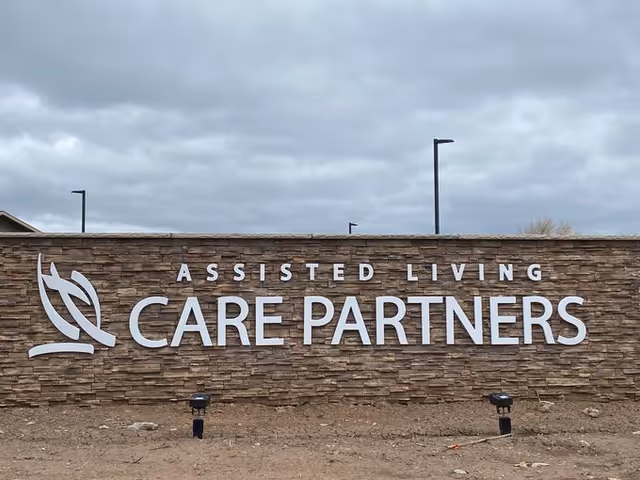 Stone wall with large white letters spelling 'ASSISTED LIVING CARE PARTNERS' and a stylized leaf logo on the left side, under a cloudy sky.