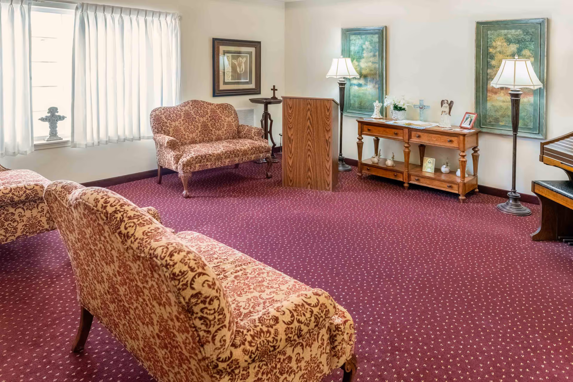 A cozy sitting room with patterned upholstered sofas on a red carpet with small white dots. The room features a wooden podium, a wooden console table with decorative items including crosses and framed pictures, two standing lamps, and two large framed paintings on the wall. A window with white curtains allows natural light into the room.