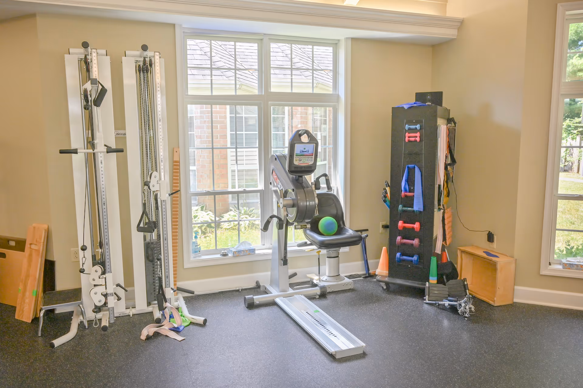 A small exercise room with various fitness equipment including a seated exercise machine with a digital display, resistance bands, small dumbbells, cones, and other workout accessories. The room has large windows letting in natural light and a view of greenery outside.