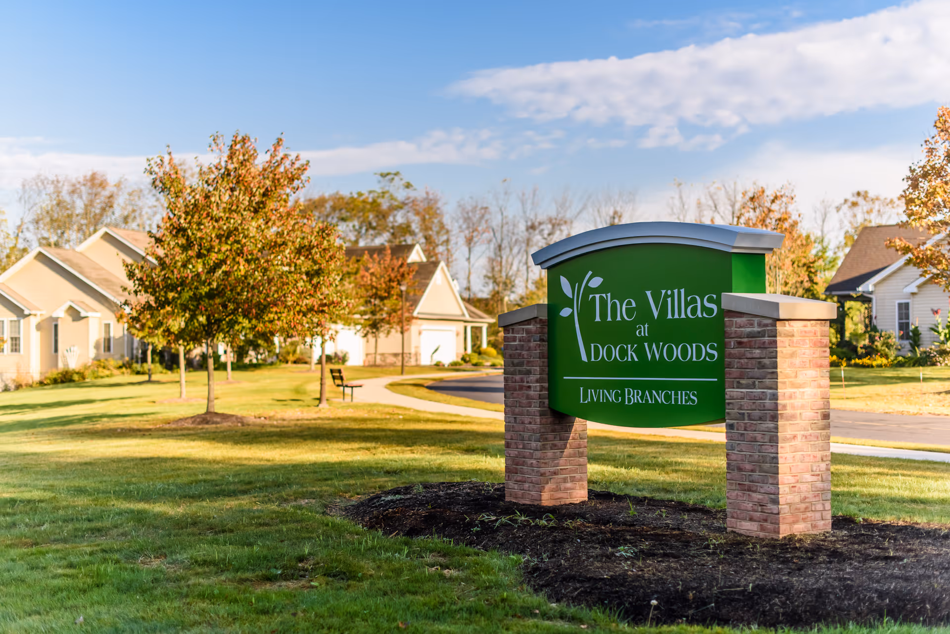 A green sign with white text reading 'The Villas at Dock Woods Living Branches' stands on a landscaped lawn with brick pillars on either side. Behind the sign, there are single-story residential buildings, trees with autumn foliage, a bench, and a paved pathway under a partly cloudy blue sky.