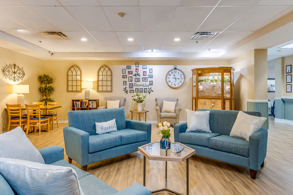 A cozy living room area in a senior living facility with three blue sofas arranged around a hexagonal glass coffee table with a vase of flowers. The walls are decorated with framed photos, a large clock, and decorative wall hangings. There is a small dining table with chairs and a lamp in the corner, and a wooden cabinet with plants on top. The room has warm lighting and a wood floor.