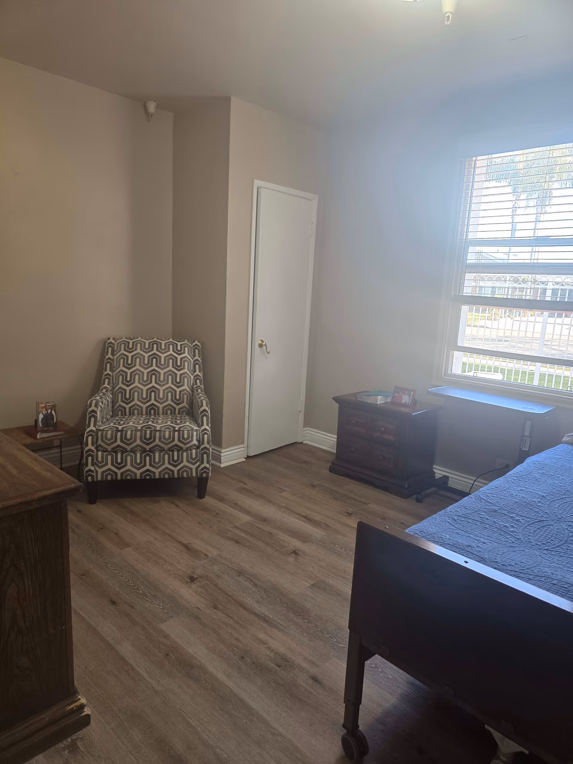 A simple bedroom with light brown walls and wood flooring. The room contains a patterned armchair, a wooden dresser, a small wooden nightstand with a framed photo and a book, and a bed with a blue quilt. A window with blinds lets in natural light.