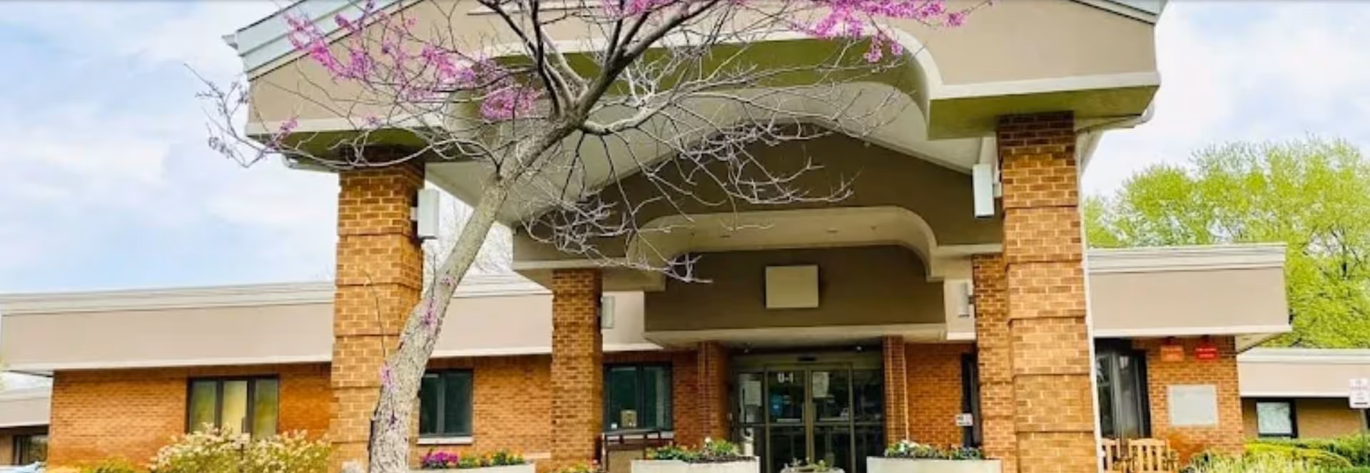 Front entrance of a brick senior living building with a covered porte-cochère and a flowering tree in front.