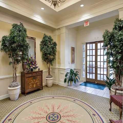 An elegant interior entryway featuring a round decorative rug with a floral pattern, two large potted trees on either side of a wooden chest with a floral arrangement and a mirror above it, a glass double door with an exit sign above, and additional potted plants near the door.