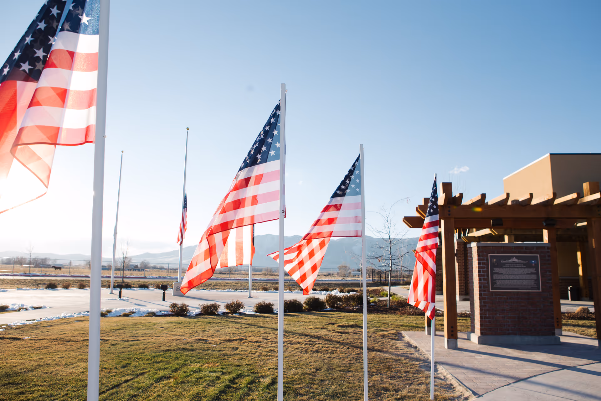 Several American flags flying on flagpoles outside a building with a pergola structure and a plaque mounted on a brick pillar, with mountains visible in the background under a clear blue sky.